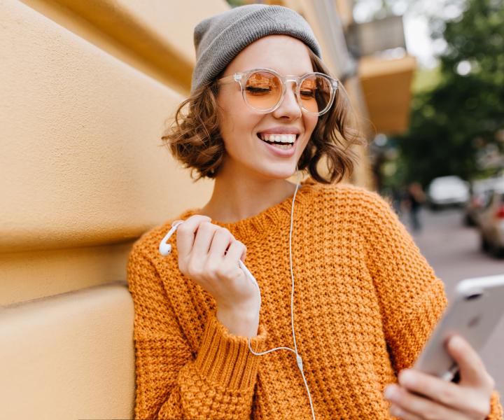 Woman in an orange sweater smiling at her smartphone while outdoors, engaging in online courses